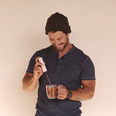 A man wearing a dark knit beanie and a navy blue polo shirt is shown from the waist up, smiling as he mixes a drink in a clear glass mug using a white handheld electric whisk.