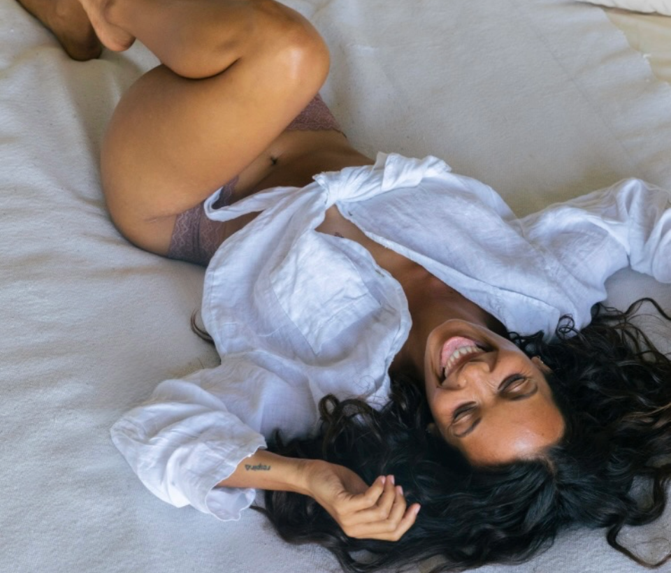 Foria supports enhanced pleasure and deeper connection. Woman in a white shirt lying on a bed. 