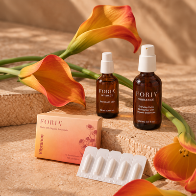 FORIV wellness products arranged on a stone surface with orange calla lilies: two amber spray bottles labeled INTHEGY and VIBRANCE, a peach box labeled Vibrance, and a blister pack of white capsules. The scene is styled with flowers and warm neutral background.