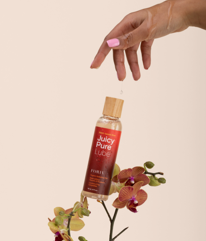 Hand holding a bottle of 'Foria Juicy Pure Lube' with an orchid on a beige. Water-based lube and oil based lube and silicone lube.