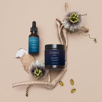 Foria Sleep Capsules CBD  including a bottle, capsules, and dropper on a beige background with flowers and driftwood.