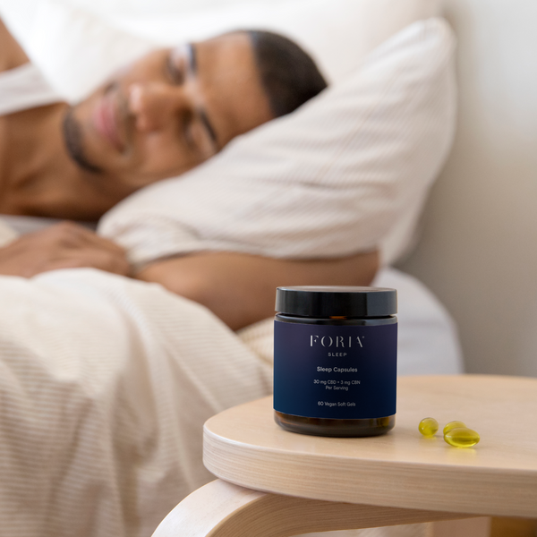 Foria Sleep Capsules CBD Person lying on a bed with Sleep supplement jar on a table in the foreground. Support deeper sleep.