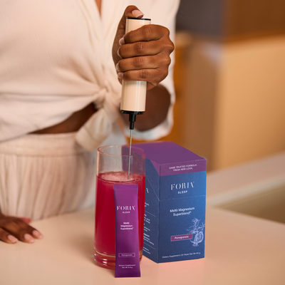 A woman in a white blouse holds a pump bottle above a glass of pink liquid, with a purple FORIV beauty box and packet on the table. The setting is a softly lit indoor space with a blurred warm background.