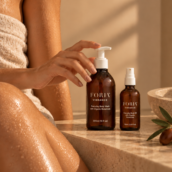 A person wrapped in a towel and leaning on a stone ledge holds a large amber pump bottle labeled FORIV VIBRANCE next to a smaller spray bottle in a warm, sunlit spa setting.