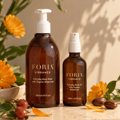 Two amber glass botanical body products from FORIA VIBRANCE, including a 300ml pump bottle of Everyday Body Wash and a 118ml spray bottle of Everyday Body Oil, styled with yellow flowers, green leaves, and argan nuts on a warm beige surface.