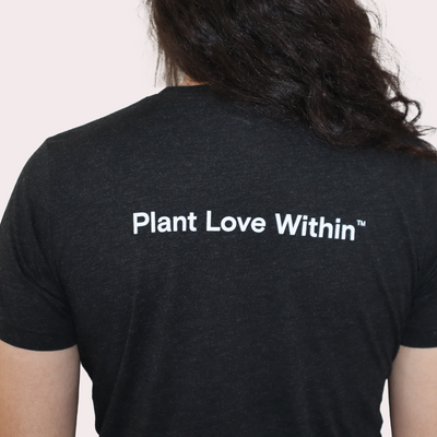 Back view of a person wearing a dark charcoal heather t-shirt with the white text 'Plant Love Within' printed across the upper back, set against a plain off-white background.